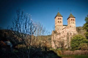 A large stone church with two square towers stands amid bare trees and greenery under a clear blue sky.