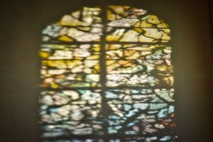 Blurry sunlight shines through an arched stained glass window, casting colorful patterns on a wall.