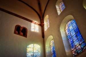 Sunlight streams through stained glass windows in a white-walled church, casting colorful patterns on the interior walls.