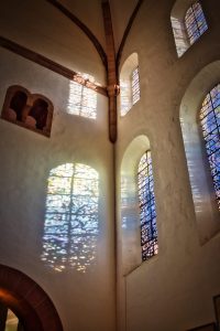 Sunlight filters through stained glass windows, casting colorful reflections on the white walls of a church interior.