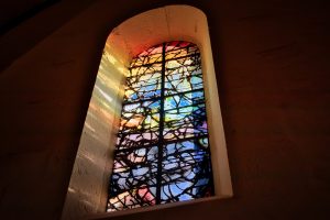 A tall, arched stained glass window set in a thick wall, illuminated by sunlight, displaying abstract patterns in blue, red, yellow, and orange tones.