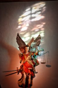 A statue of an angel with wings, holding a sword and scales, stands against a white wall with colored light reflected above it.