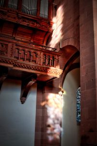 Sunlight filters through a stained glass window, casting colored reflections on the stone wall beneath a wooden balcony inside a church.