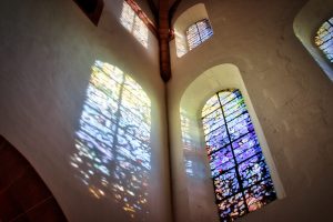 Colorful sunlight filters through stained glass windows, casting vibrant patterns on the white stone walls of a church interior.