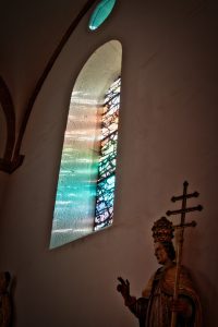 A stained glass window casts colorful light onto a white wall inside a church, with a statue of a religious figure holding a staff in the foreground.
