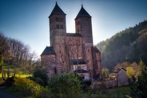 A stone church with two tall towers stands amid trees and greenery under a clear blue sky.