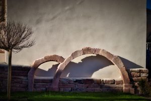 A white wall with two partial stone arches and a leafless tree in front, casting shadows on a grassy area.