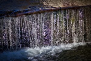 Close-up of water cascading over a stone ledge, creating a small waterfall with visible white foam and sunlight reflecting off the flowing water.