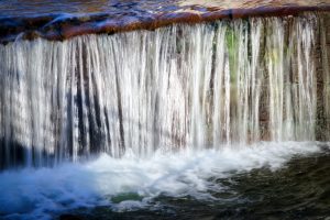 Water cascades in thin streams over a ledge, creating a vertical waterfall with white foam forming at the base.