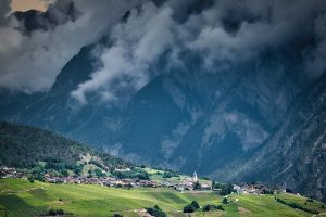 A small village with a church is nestled among green fields at the foot of tall mountains, partially covered by low-hanging clouds.