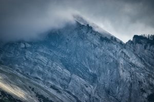 A rugged, rocky mountain slope with scattered trees is partially covered by low-hanging clouds under an overcast sky.