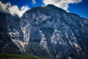 Steep, rocky mountain slopes rise sharply under a blue sky with scattered clouds, partially covered with green forest at the base.