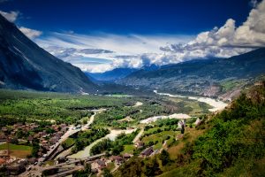 A wide valley with a river running through forests and a village, surrounded by mountains under a partly cloudy blue sky.