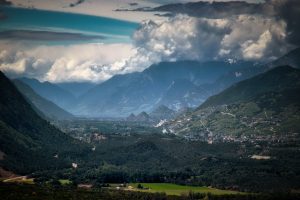 View of a lush valley with scattered towns, surrounded by forested hills and dramatic mountains under a cloudy sky.