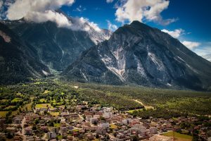 A small town with clustered buildings sits at the base of tall, forested mountains under a partly cloudy sky.