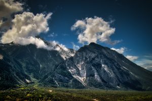 Mountain range with steep rocky slopes and scattered clouds under a deep blue sky, with green forested areas at the base.