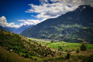 A lush green valley with scattered houses lies beneath tall, forested mountains under a partly cloudy sky.