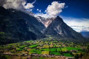 A green valley with scattered buildings lies beneath tall, rugged mountains under a partly cloudy blue sky.