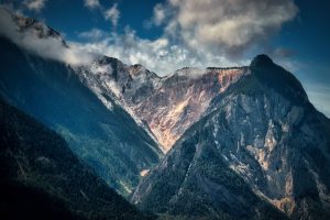 Clouds partially cover rugged, forested mountains with a sunlit, rocky slope in the center under a blue sky.