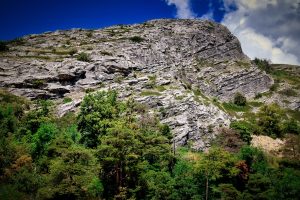 Large rocky hillside with exposed gray stone and green trees at the base, under a partly cloudy blue sky.