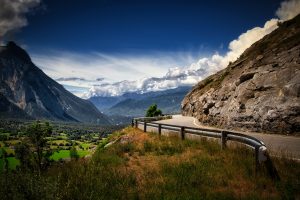 A winding mountain road curves along a rocky hillside, overlooking a green valley with distant mountains under a partly cloudy sky.