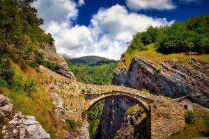 Stone arch bridge spans a deep rocky gorge, surrounded by green hills and trees under a partly cloudy blue sky. A small white building sits near the bridge.