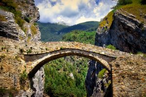 Stone arch bridge spans a rocky gorge with green forested hills and a partly cloudy sky in the background.