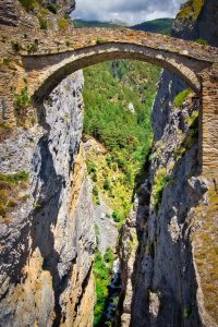 A stone arch bridge spans a deep, narrow canyon with steep rocky cliffs and dense green forest below.