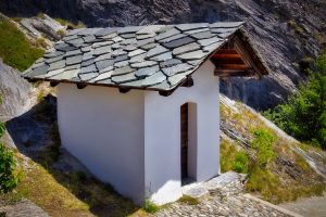 Small white building with a stone-tiled roof and wooden door, situated on a hillside with rocky terrain and some greenery in the background.