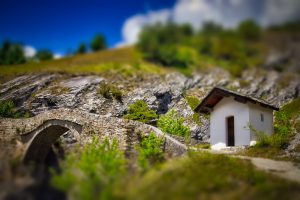 A small white house with a wooden roof stands near a stone bridge on a hillside with greenery under a blue sky.