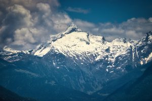 Snow-capped mountain peaks under a partly cloudy sky, with forested lower slopes and rugged terrain.