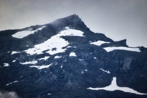 Rocky mountain peak with patches of snow under a cloudy sky. A small cross is visible at the summit.