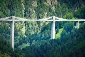 A tall concrete bridge with multiple supports spans a lush, green forested valley with steep hillsides in the background.