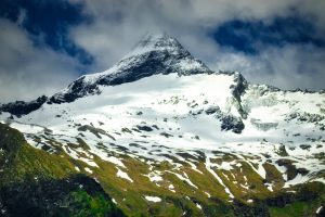 Snow-covered mountain peak partially obscured by clouds, with green and rocky slopes in the foreground under a partly cloudy sky.