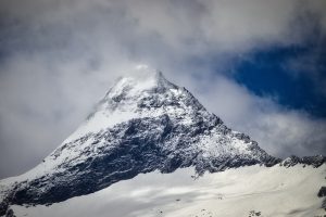 Snow-covered mountain peak partially obscured by clouds, with blue sky visible in the background.