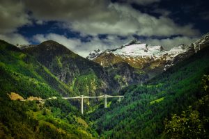A tall concrete bridge spans a lush green valley with forested hills and snow-capped mountains in the background under a cloudy sky.