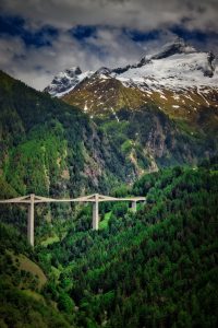 A concrete bridge spans a forested valley with snow-capped mountains in the background under a partly cloudy sky.