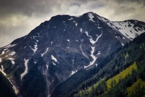 A rocky mountain peak with patches of snow and forested slopes under a cloudy sky.