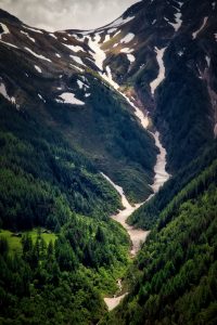 Mountain slope with patches of snow, dense green forest, and a winding path leading through the valley.