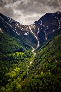 A lush green valley with dense trees lies between two tall mountains, with patches of snow near the peaks under a cloudy sky.