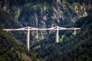 A long concrete bridge with tall supports spans a forested valley, surrounded by dense green trees and rugged hills.