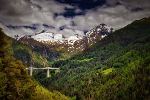 A concrete bridge spans a forested mountain valley with snow-capped peaks in the background under a partly cloudy sky.