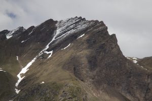 A rugged mountain peak with patches of snow and green slopes under a cloudy sky.