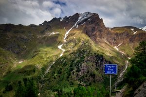Mountain scenery with patches of snow, green slopes, and a blue sign in the foreground reading "Simplon Pass 2005 m.