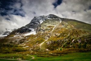 Mountain landscape with partly snow-covered rocky peak, scattered green vegetation, a winding path, and a cloudy sky overhead.